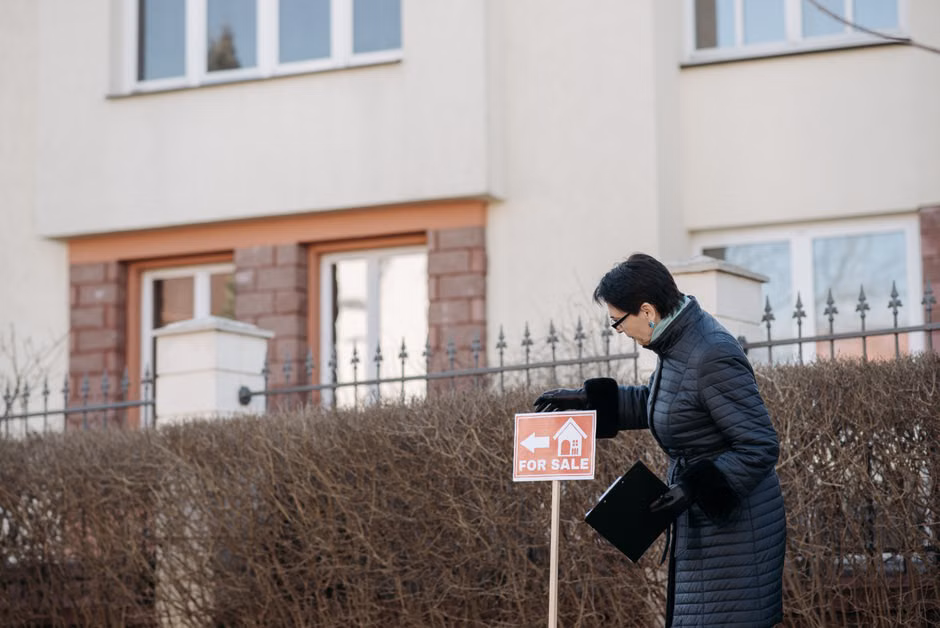 Real estate agent in a black coat placing a 'For Sale' sign in front of a house.